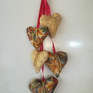 Op de afbeelding: Een reeks van vier stoffen harten met gouden kralenaccenten en een rood lint dat aan een wit oppervlak hangt. De harten zijn versierd met een gouden en bruin bloemenpatroon.