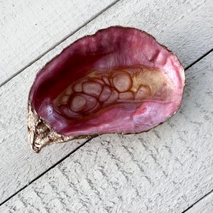 May include: A decorative oyster shell with a gold-painted edge and a pink and gold interior. The shell is resting on a white wooden surface. This unique piece can be used as a jewelry dish or a decorative accent.