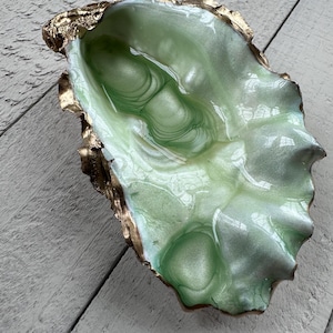 May include: A decorative oyster shell dish featuring a glossy, pale green and white marbled interior finish with a metallic gold painted edge. The shell has an irregular, organic shape with a smooth, reflective surface and a textured, gilded rim.
