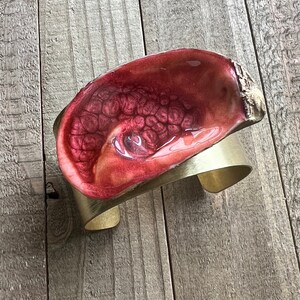 May include: A red resin bracelet with a gold metal band. The resin has a marbled effect with a glossy finish.