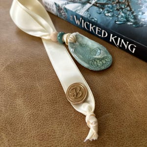 May include: Ivory ribbon bookmark with a teal and white pendant, a wooden bead, and a gold-colored seal with the letter "B". The bookmark is next to a book titled "The Wicked King".