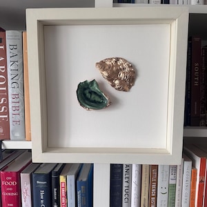 May include: A framed artwork featuring two oyster shell sculptures. One shell is gold-colored, and the other is green and gold. The artwork is displayed against a white background within a white frame. The artwork is displayed on a shelf with books.