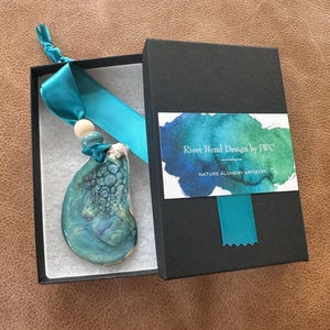 May include: A teal and blue pendant with a marbled design, presented in a black gift box with a teal ribbon. The box has a label that reads "River Bend Design by JWC" and "NATURE ALCHEMY ARTISTRY."