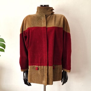Peut inclure: Une veste en velours côtelé marron et rouge avec un col montant et une fermeture boutonnée. La veste présente un design bicolore contrasté avec un haut marron et un bas rouge.