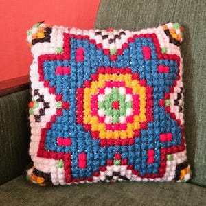 May include: Square, handmade pillow with a colorful geometric design. The pillow features a central pattern in blue, yellow, red, and green, surrounded by a white border. The pillow is placed on a green chair.