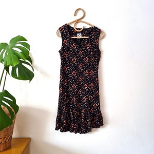 K&ouml;nnte beinhalten: Ein schwarzes, &auml;rmelloses Kleid mit einem Blumendruck in Orange- und Wei&szlig;t&ouml;nen. Das Kleid hat einen R&uuml;schen-Saum und einen runden Ausschnitt.