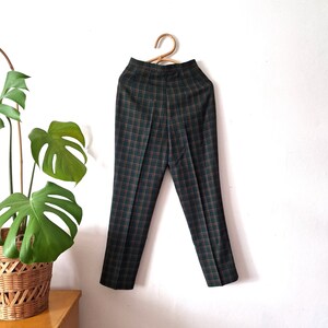May include: Green and brown plaid pants hanging on a wooden hanger against a white wall.