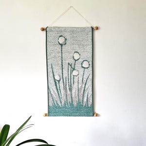 Könnte beinhalten: Ein rechteckiger, gewebter Wandbehang mit einem stilisierten botanischen Muster aus weißen, löwenzahnartigen Blüten auf langen Stielen vor einem strukturierten hellgrauen und dunkel-türkisfarbenen Hintergrund, aufgehängt an einem Holzstab.