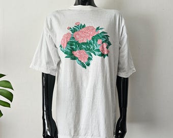 Puede incluir: Una camiseta gris claro con un diseño floral. El estampado gráfico muestra flores rosas y hojas verdes. La camiseta se exhibe en un maniquí negro sobre un fondo blanco.