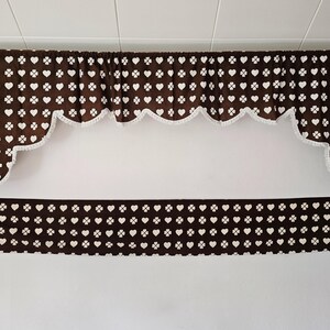 May include: A brown window valance featuring a repeating white pattern of hearts and four-leaf clovers. The fabric has a gathered top and a scalloped bottom edge trimmed with white fringe. A matching straight brown fabric strip is positioned below.