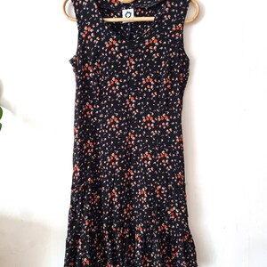K&ouml;nnte beinhalten: Ein &auml;rmelloses schwarzes Kleid mit einem Blumendruck in Orange-, Gelb- und Wei&szlig;t&ouml;nen. Das Kleid hat einen R&uuml;schen-Saum.