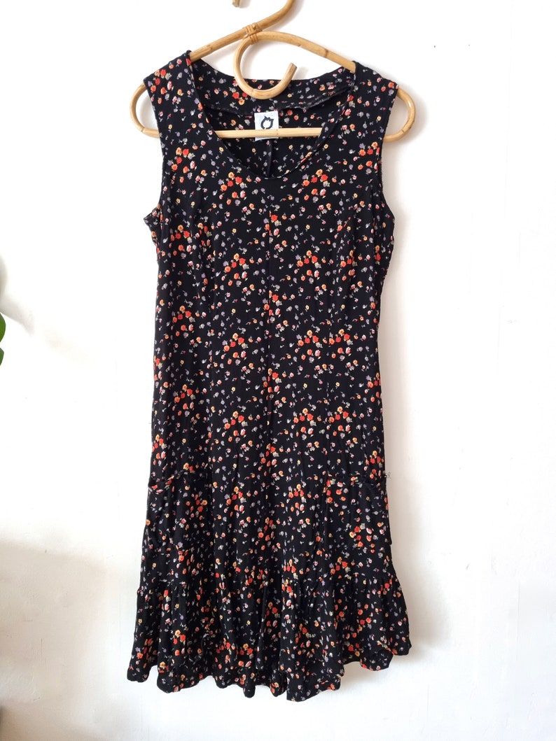 K&ouml;nnte beinhalten: Ein &auml;rmelloses schwarzes Kleid mit einem Blumendruck in Orange-, Gelb- und Wei&szlig;t&ouml;nen. Das Kleid hat einen R&uuml;schen-Saum.
