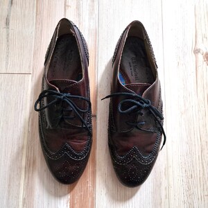 Pu&ograve; includere: Un paio di scarpe eleganti in pelle bordeaux scuro con lacci neri. Le scarpe hanno un design a punta alata con perforazioni decorative e sono appoggiate su una superficie di legno chiaro. Il marchio "C.B. Mariani" &egrave; visibile all'interno.