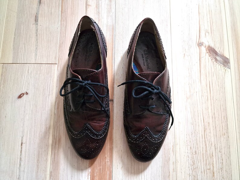 Pu&ograve; includere: Un paio di scarpe eleganti in pelle bordeaux scuro con lacci neri. Le scarpe hanno un design a punta alata con perforazioni decorative e sono appoggiate su una superficie di legno chiaro. Il marchio "C.B. Mariani" &egrave; visibile all'interno.