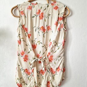 May include: A sleeveless, cream-colored blouse with a floral print featuring pink flowers and green leaves. The blouse has a tie at the back.