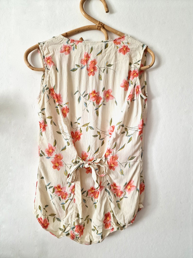 May include: A sleeveless, cream-colored blouse with a floral print featuring pink flowers and green leaves. The blouse has a tie at the back.