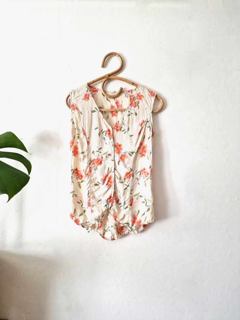 May include: A sleeveless white blouse with a floral print featuring pink flowers and green leaves. The blouse has a button-up front and a gathered waist.
