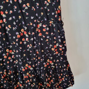 K&ouml;nnte beinhalten: Ein schwarzes Kleid mit Blumenmuster mit kleinen roten, orangefarbenen und violetten Bl&uuml;ten. Das Kleid hat einen R&uuml;schen-Saum und eine Tasche an der Seite.