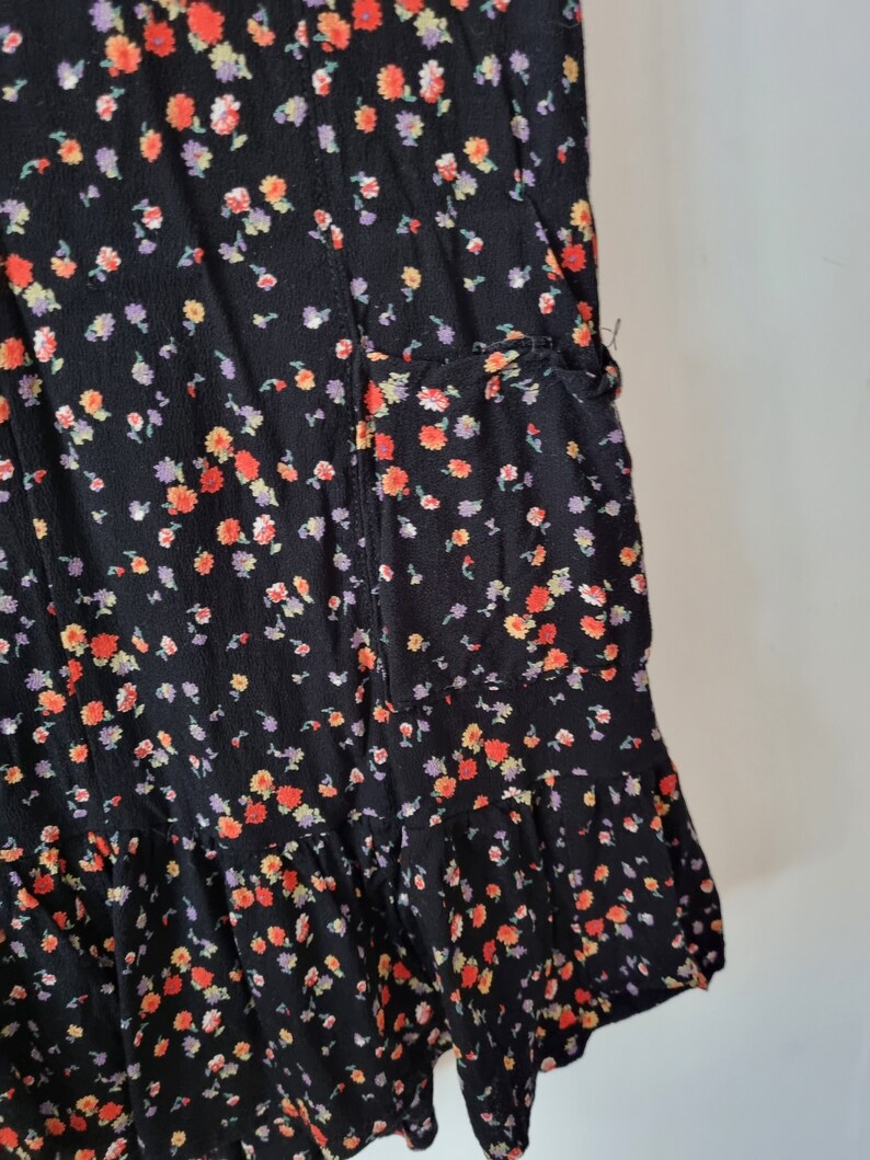 K&ouml;nnte beinhalten: Ein schwarzes Kleid mit Blumenmuster mit kleinen roten, orangefarbenen und violetten Bl&uuml;ten. Das Kleid hat einen R&uuml;schen-Saum und eine Tasche an der Seite.