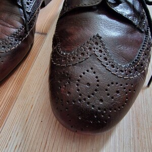 Pu&ograve; includere: Primo piano di due scarpe Oxford in pelle marrone con perforazioni decorative e dettagli wingtip. Le scarpe hanno lacci neri e sono appoggiate su un pavimento in legno chiaro.