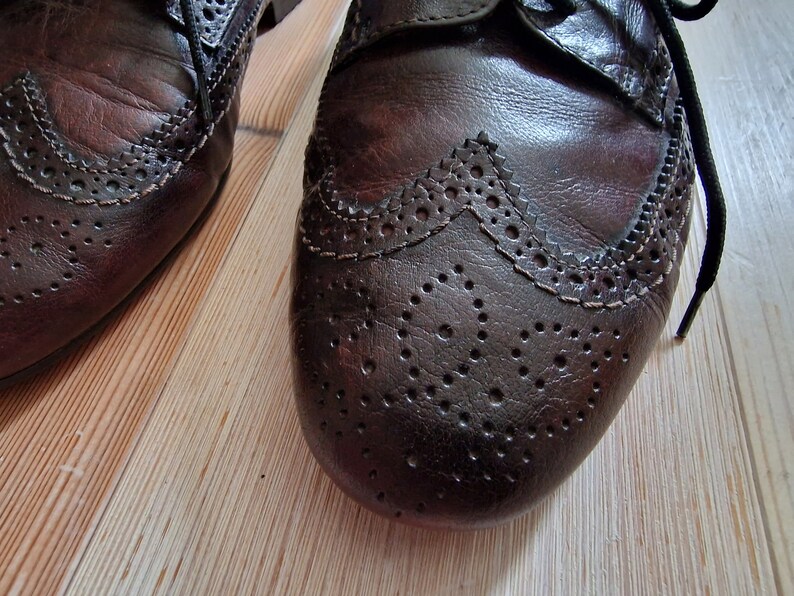 Pu&ograve; includere: Primo piano di due scarpe Oxford in pelle marrone con perforazioni decorative e dettagli wingtip. Le scarpe hanno lacci neri e sono appoggiate su un pavimento in legno chiaro.