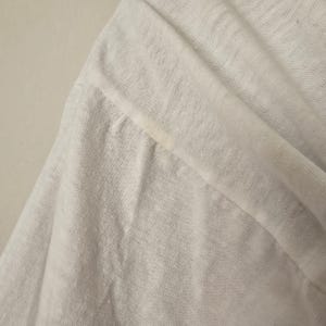 Op de afbeelding: Close-up van een witte, lichte stof, mogelijk linnen of een vergelijkbaar ademend materiaal. De stof is gevouwen en toont de textuur en de drapering. Er zit een kleine vlek op de stof. De afbeelding benadrukt de weefsel en de kleur van het materiaal.