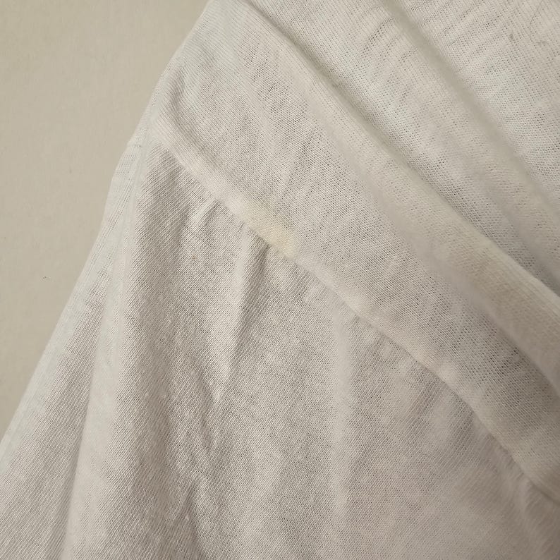 Op de afbeelding: Close-up van een witte, lichte stof, mogelijk linnen of een vergelijkbaar ademend materiaal. De stof is gevouwen en toont de textuur en de drapering. Er zit een kleine vlek op de stof. De afbeelding benadrukt de weefsel en de kleur van het materiaal.