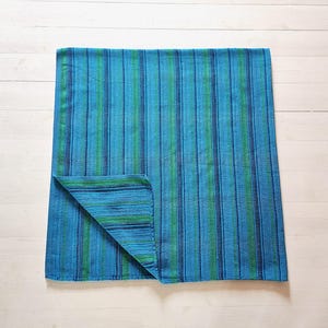 Könnte beinhalten: Ein rechteckiges Textil mit einem vertikalen Streifenmuster in den Farben leuchtendes Blau, Dunkelblau und kräftiges Grün. Der Stoff hat ein strukturiertes Gewebe und eine Ecke ist umgeschlagen, um das gleiche Streifenmuster auf der Rückseite zu zeigen.