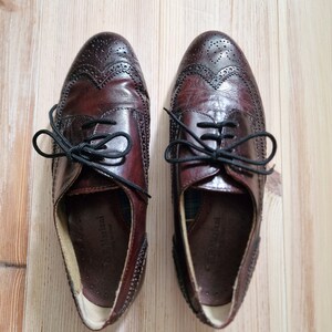 Pu&ograve; includere: Un paio di scarpe Oxford in pelle bordeaux con lacci neri. Le scarpe presentano perforazioni decorative e un classico design a punta di ala. Le scarpe sono esposte su una superficie di legno chiaro.