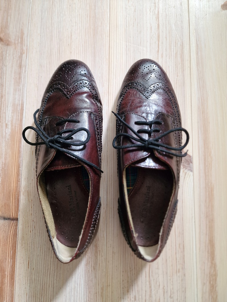 Pu&ograve; includere: Un paio di scarpe Oxford in pelle bordeaux con lacci neri. Le scarpe presentano perforazioni decorative e un classico design a punta di ala. Le scarpe sono esposte su una superficie di legno chiaro.