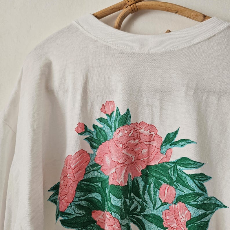 Op de afbeelding: Wit T-shirt met een bloemenontwerp van roze pioenrozen en groene bladeren. De bloemen zijn gegroepeerd, waardoor een levendig ontwerp ontstaat. Het shirt hangt aan een houten hanger.