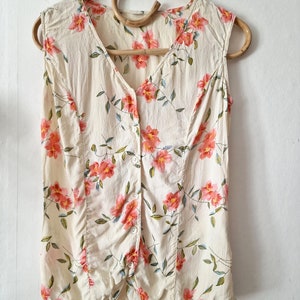 May include: A sleeveless, cream-colored blouse with a floral print featuring pink flowers and green leaves. The blouse has a button-up front and a V-neck.