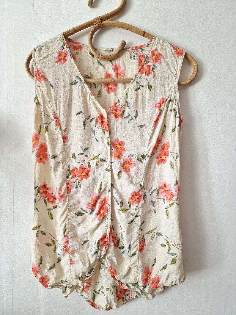May include: A sleeveless, cream-colored blouse with a floral print featuring pink flowers and green leaves. The blouse has a button-up front and a V-neck.