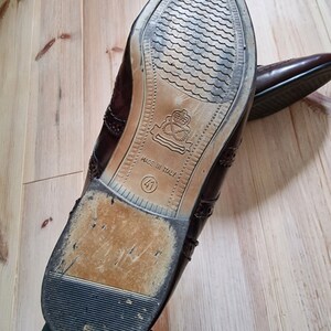 Pu&ograve; includere: Un paio di scarpe eleganti in pelle marrone con decorazioni traforate. Le suole sono visibili, con un design strutturato e la scritta "MADE IN ITALY" e la taglia 41. Le scarpe sono su una superficie di legno chiaro.