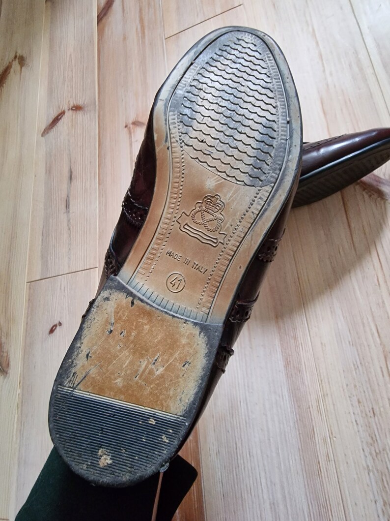 Pu&ograve; includere: Un paio di scarpe eleganti in pelle marrone con decorazioni traforate. Le suole sono visibili, con un design strutturato e la scritta "MADE IN ITALY" e la taglia 41. Le scarpe sono su una superficie di legno chiaro.