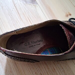 Pu&ograve; includere: Primo piano di una scarpa in pelle marrone con cuciture decorative e lacci neri. L'interno della scarpa presenta un motivo a quadri e il testo "C.B. Mariani MADE IN ITALY".