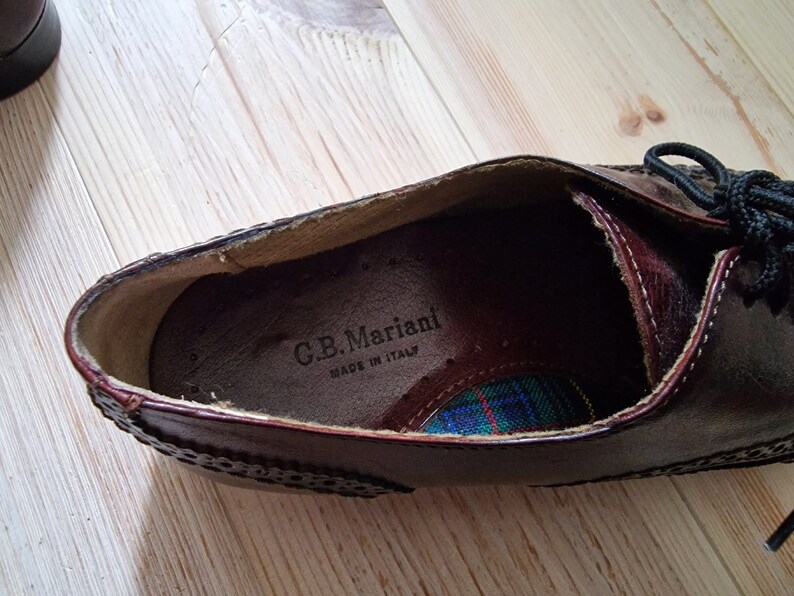 Pu&ograve; includere: Primo piano di una scarpa in pelle marrone con cuciture decorative e lacci neri. L'interno della scarpa presenta un motivo a quadri e il testo "C.B. Mariani MADE IN ITALY".