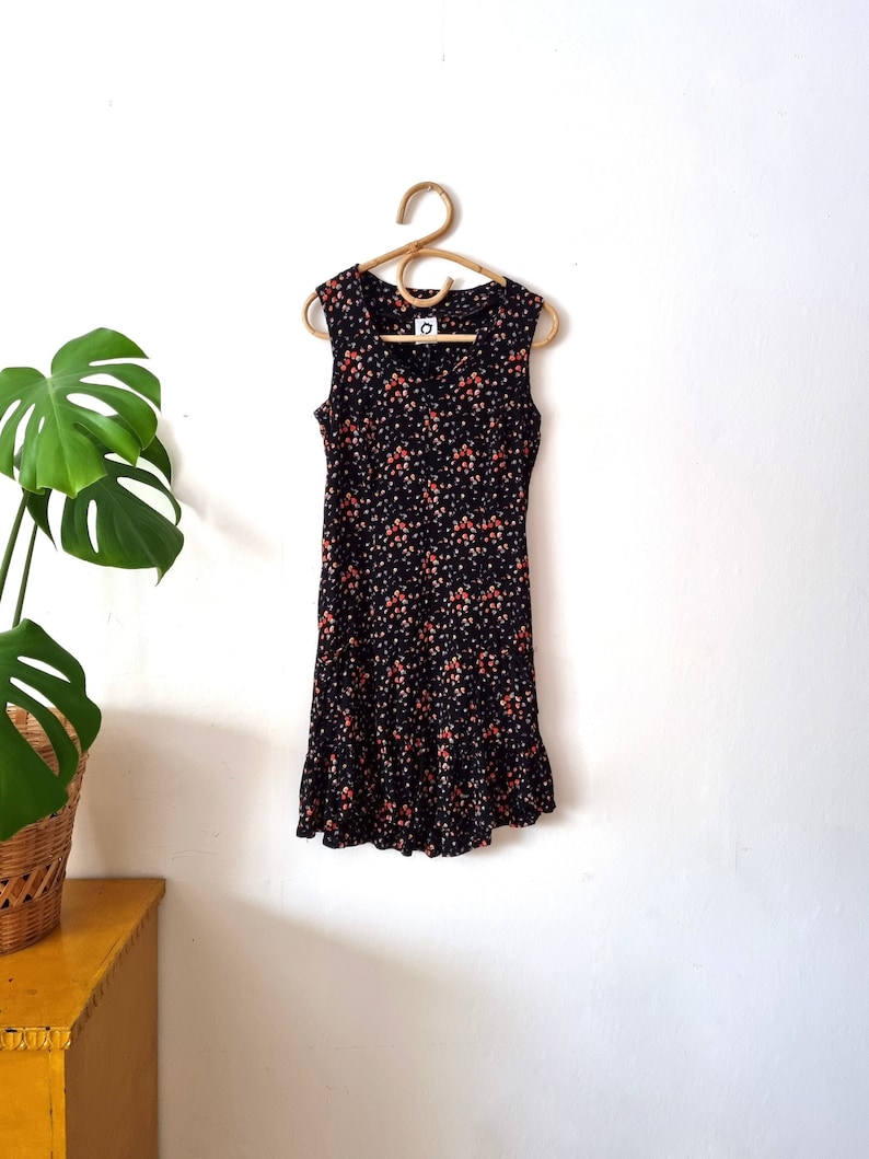 K&ouml;nnte beinhalten: Ein schwarzes, &auml;rmelloses Kleid mit einem Blumendruck in Orange- und Wei&szlig;t&ouml;nen. Das Kleid hat einen R&uuml;schen-Saum und einen runden Ausschnitt.