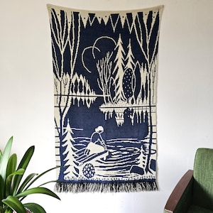 May include: A rectangular woven wall hanging featuring a dark blue and cream-colored landscape scene. The design depicts a person kneeling on a wooden dock by a lake, surrounded by tall pine trees and forest foliage, finished with a fringed bottom edge.