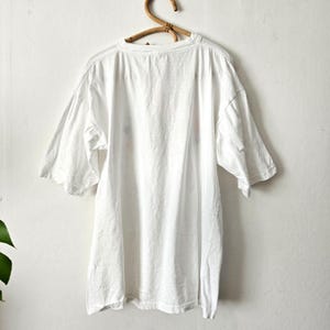 Op de afbeelding: Een wit, oversized t-shirt met korte mouwen hangt aan een houten hanger. Het shirt lijkt gemaakt van een zachte, lichte stof. De achtergrond is een effen witte muur.