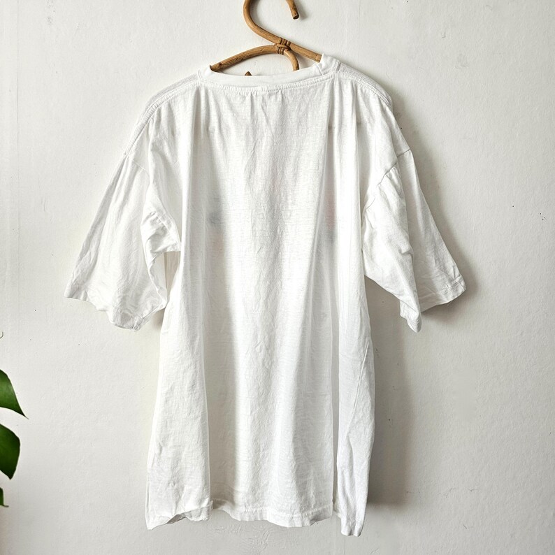 Op de afbeelding: Een wit, oversized t-shirt met korte mouwen hangt aan een houten hanger. Het shirt lijkt gemaakt van een zachte, lichte stof. De achtergrond is een effen witte muur.