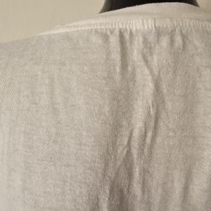 Op de afbeelding: Close-up van een wit t-shirt, met de achterkant van de nek en schouderpartij. De stof heeft een licht getextureerde uitstraling met subtiele kleurvariaties. Het shirt staat op een zwarte mannequin.
