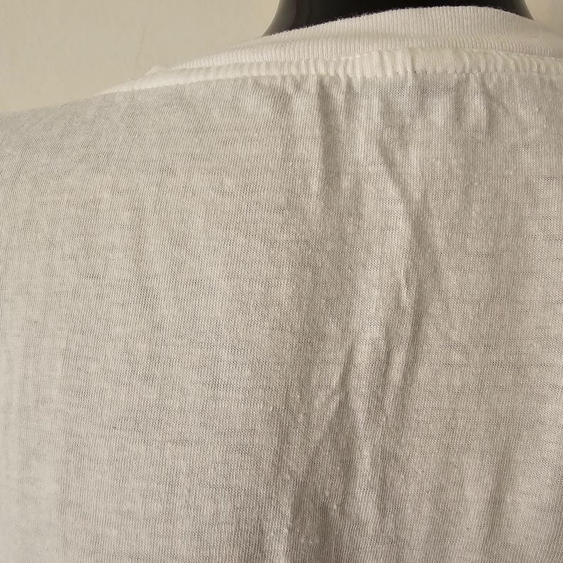 Op de afbeelding: Close-up van een wit t-shirt, met de achterkant van de nek en schouderpartij. De stof heeft een licht getextureerde uitstraling met subtiele kleurvariaties. Het shirt staat op een zwarte mannequin.