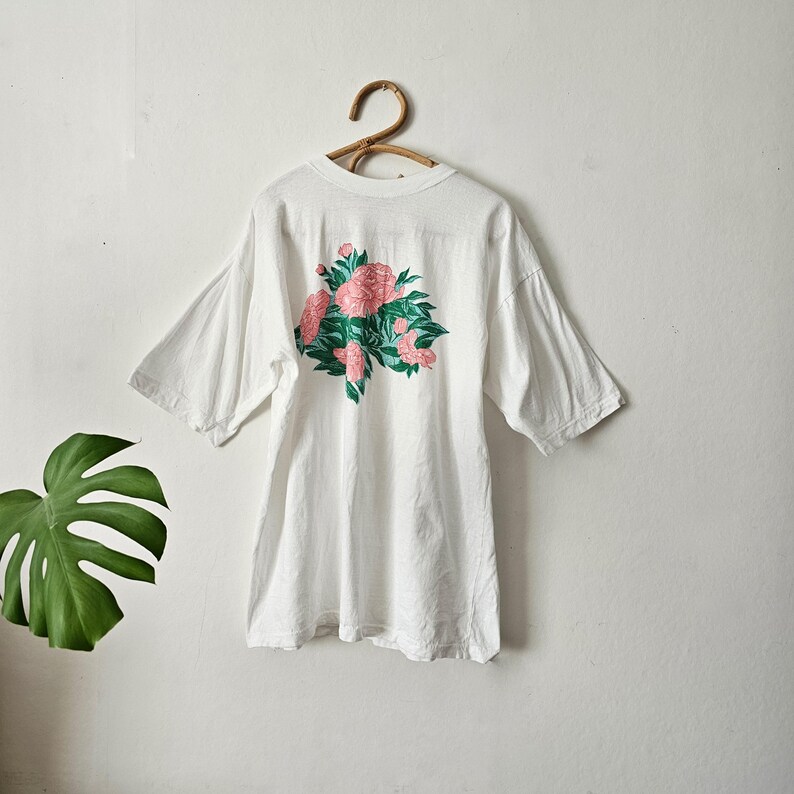 Op de afbeelding: Wit T-shirt met een bloemenontwerp. Het shirt heeft een groot roze en groen bloemenboeket op de achterkant. Het shirt met korte mouwen hangt aan een houten hanger tegen een witte muur.