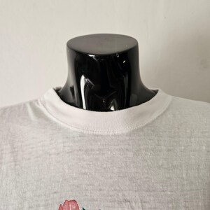 Op de afbeelding: Een grijs gem&ecirc;leerd T-shirt met een witte ronde hals. De shirt heeft een bloemenontwerp aan de onderkant. Het T-shirt wordt getoond op een zwarte mannequinhoofd tegen een witte achtergrond.