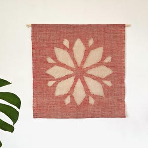 Puede incluir: Un tapiz de pared tejido en rojo y crema. El textil cuadrado presenta un gran diseño floral simétrico en crema sobre un fondo rojo. El tapiz de pared está montado en una clavija de madera para su exhibición.