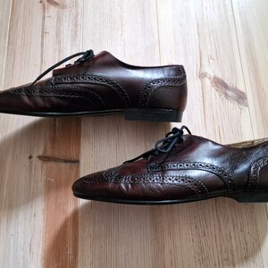 Pu&ograve; includere: Un paio di scarpe Oxford in pelle marrone scuro con perforazioni decorative e lacci neri. Le scarpe sono esposte su una superficie di legno marrone chiaro.