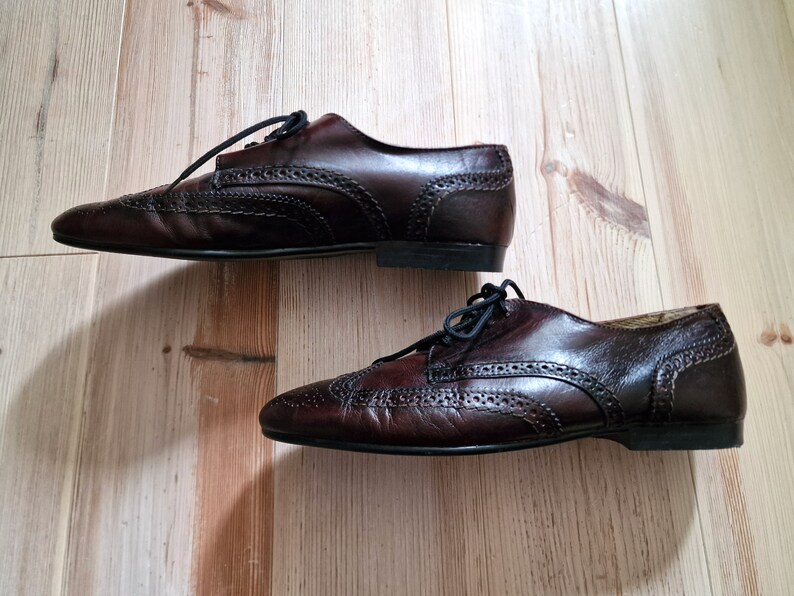 Pu&ograve; includere: Un paio di scarpe Oxford in pelle marrone scuro con perforazioni decorative e lacci neri. Le scarpe sono esposte su una superficie di legno marrone chiaro.