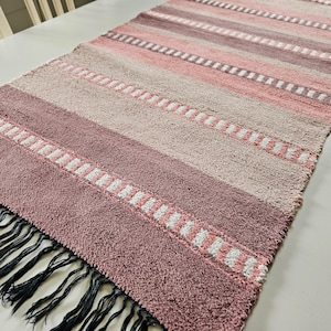 Op de afbeelding: Een rechthoekige tafelloper met een geweven streeppatroon in tinten oudroze, mauve en beige. De stof heeft een getextureerd oppervlak met dunne witte horizontale accenten tussen de strepen en zwarte franjes langs de korte zijde.