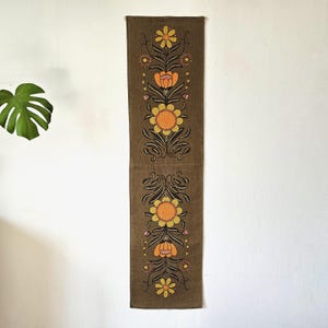 Può includere: Un lungo arazzo da parete rettangolare marrone con un motivo floreale. Il disegno presenta fiori gialli e arancioni con steli e foglie nere. L'arazzo è realizzato in tessuto strutturato ed è appeso a una parete bianca.